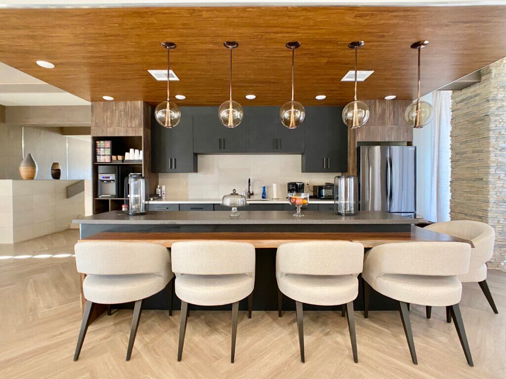 Four cushioned chairs sit at a wood table under lighting fixtures, an equipped kitchen in the background